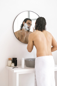 Shirtless young man with white towel on hips applying shaving foam on his beard in front of mirror in the bathroom