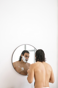 Shirtless young man applying shaving foam on his beard while standing in front of mirror in the bathroom