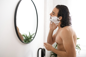 Shirtless young muscular man applying shaving foam on face while looking in mirror in the bathroom