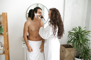Young long-haired woman in white bathrobe applying shaving foam on beard of her husband while standing in front of him in bathroom