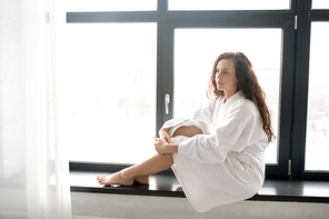 Young brunette serene woman in soft white bathrobe relaxing on windowsill on weekend morning at home