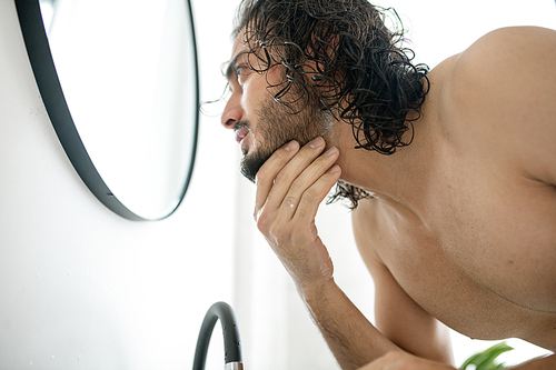 Young shirtless man bending over sink in front of mirror while touching his beard before shaving it