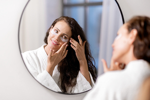 Happy young woman in white bathrobe standing in front of mirror in bathroom and putting undereye patches on face