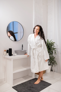 Pretty young smiling brunette female in white bathrobe standing on small rug in front of camera in bathroom