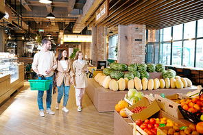 Casual young family of three moving down large contemporary supermarket while passing by fresh fruits
