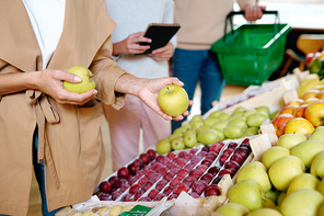 Young woman in beige trenchcoat holding two ripe granny smith apples while choosing some on fruit display in supermarket