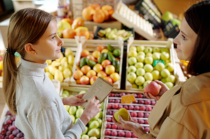 Young brunette woman with apples looking at her daughter with shopping list while choosing fruits in supermarket