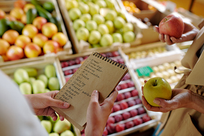 Hands of girl holding notepad with shopping list by display with plums, pears and peaches while visiting supermarket with mother