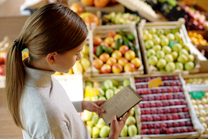 Pretty youthful girl in casual pullover looking through shopping list in notepad while standing by fruit display in supermarket