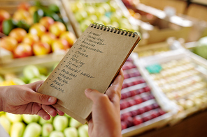 Notepad with list of products to buy in supermarket held by youthful girl standing by display with ripe fresh fruits
