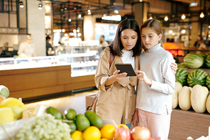 Affectionate young woman and her daughter holding notepad with shopping list while reading it by fruit display in supermarket