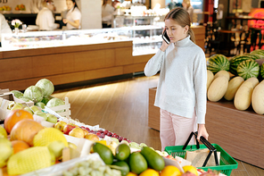 Pretty girl in casualwear talking to her mom on mobile phone while standing by fruit display in contemporary supermarket