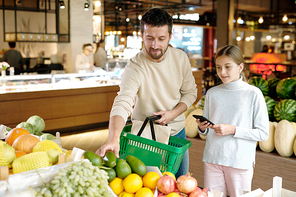 Young man with basket and his daughter choosing avocado and other fruits while visiting large modern supermarket