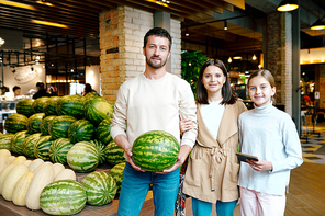 Young casual family of three buying ripe watermelon while standing in large supermarket by fruit display