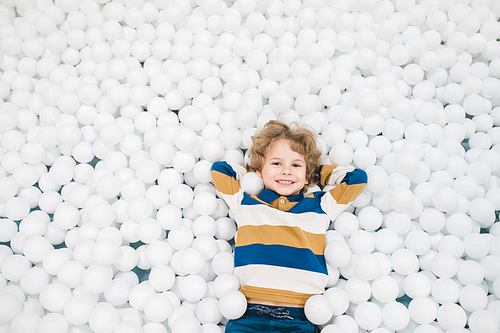 Adorable joyful little boy in casualwear lying among white balloons and looking at you while having fun