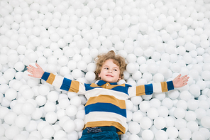 Cute little boy in casualwear outstretching his arms while lying among white balloons and enjoying play in isolation