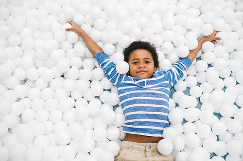 Cute little African boy in casualwear raising arms while having fun among white balloons during play in isolation