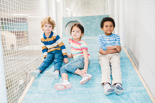 Group of three adorable intercultural children looking at you while sitting on play area at leisure center