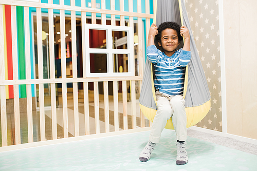 Happy little African boy in casualwear sitting in hammock hanging from ceiling on play area of leisure center