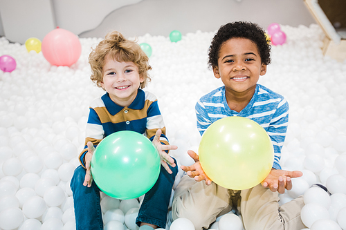 Cute cheerful intercultural little boys in striped shirts playing with green and yellow balloons in children room or kindergarten