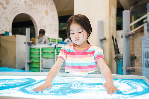 Cute little girl of Asian ethnicity making picture of blue sand by her hands while spending time in children room