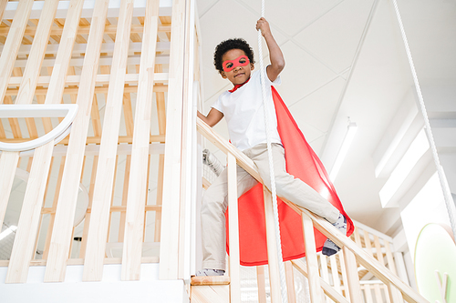 Adorable boy of African ethnicity wearing red mantle of superman holding by rope while gliding down railings