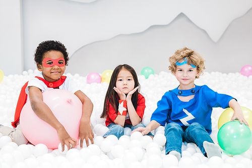 Adorable little intercultural friends in costumes playing with white balloons in children room or kindergarten