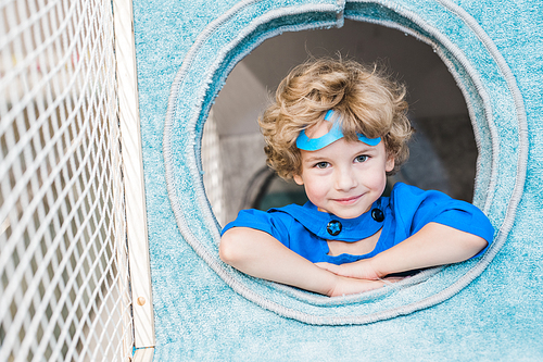 Blonde curly little boy in blue costume of superman looking at you while playing in soft house on play area