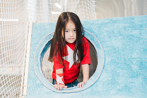 Cute little girl of Asian ethnicity in costume of super girl looking at you while playing in soft house on play area