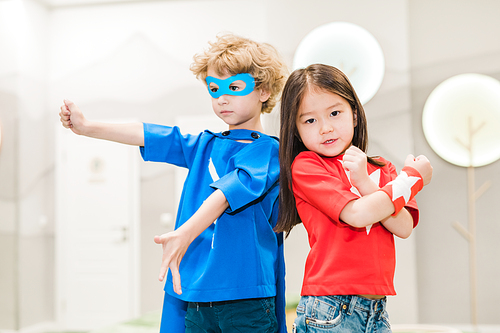 Two intercultural children in costumes of super heroes standing close to each other in front of camera during play