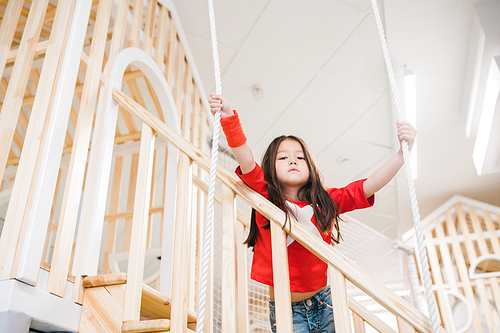 Cute little Asian girl looking at you while standing on wooden staircase and holding by ropes on playground