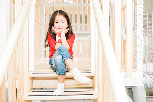 Adorable Asian girl in casualwear sitting on wooden stairs at play area between railings and looking at you