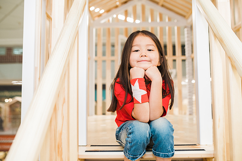 Pretty restful little girl in blue jeans and red t-shirt sitting on wooden stairs at play area between railings