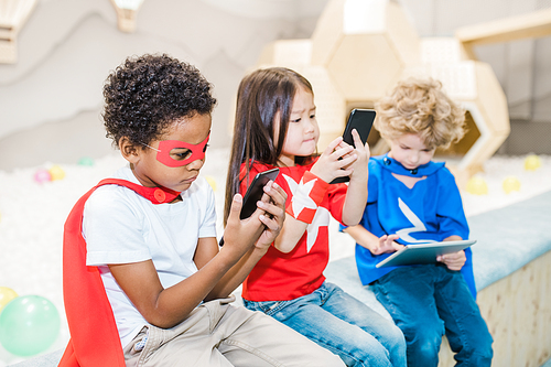 Cute African boy in costume of superhero and his two friends using gadgets at leisure while playing games or watching cartoons