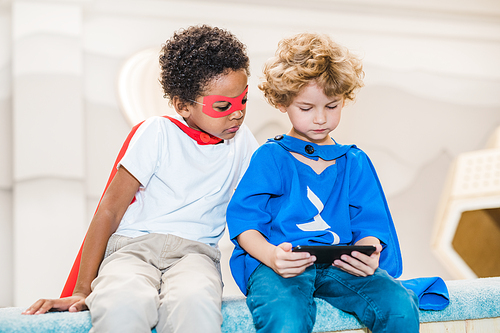 Adorable blonde boy and his African friend in costumes of superman watching curious stuff in smartphone after playing