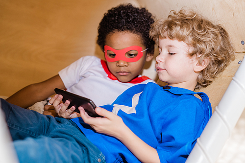 Two little intercultural restful boys in costumes of superman looking at screen of smartphone while relaxing at leisure