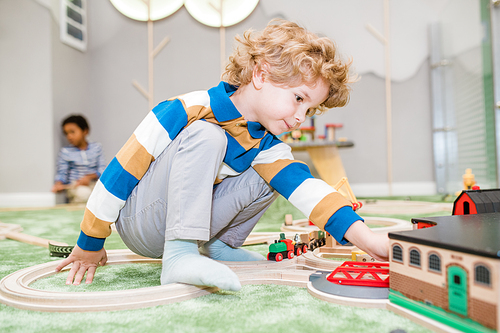 Adorable blond little boy in casualwear playing on the floor with toy trains and house in kindergarten or children center