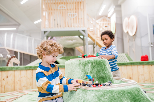 Two adorable little boys in casualwear playing toy trains together on green play area in kindergarten or leisure center