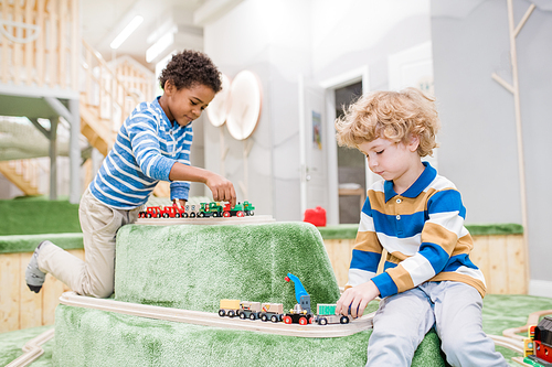 Two intercultural little boys sitting on playground while playing toy trains together in contemporary children center