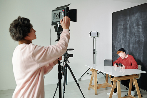 Young man in casualwear preparing video camera before shooting while standing in studio in front of male vlogger