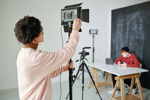 Young cameraman in casualwear preparing video shooting equipment while standing in studio in front of male vlogger