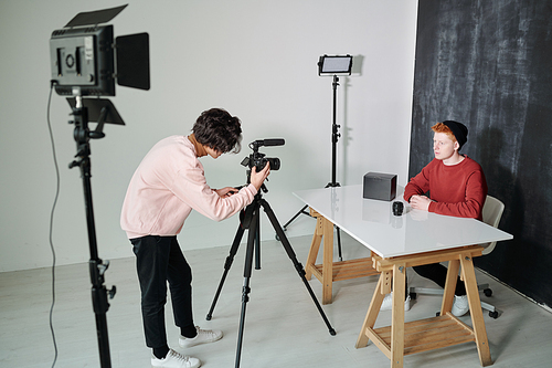 Young cameraman bending in front of video shooting equipment while standing in studio in front of male vlogger
