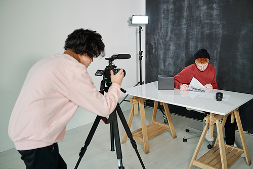 Guy in casualwear bending in front of video camera while shooting male vlogger sitting by desk in studio