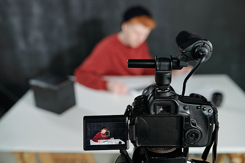Screen of video camera in front of young male vlogger sitting by desk against black background in studio