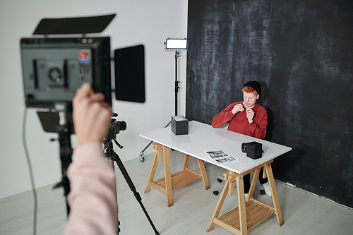 Young vlogger in casualwear sitting by desk against black background in studio and preparing for video shooting