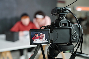 Two young contemporary male vloggers on screen of video camera standing in front of desk in studio