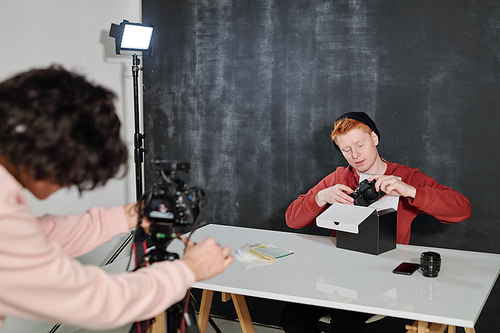 Young operator by video camera shooting male vlogger unpacking box and taking out new photocamera while sitting by desk in studio