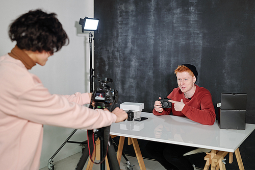 Young operator shooting happy male vlogger pointing at new photocamera while showing it to online audience in studio