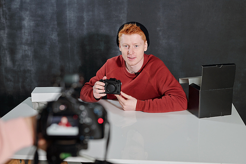 Young man in casualwear sitting by desk against black background in studio and talking about new photocamera