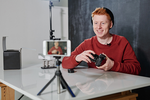 Young cheerful man in casualwear sitting by desk in front of video camera in studio and presenting his new photocamera
